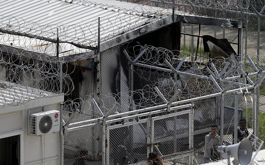 FILE - In this photo taken on Thursday, March 16, 2017 migrants stand behind a fence at the Moria refugee detention center on the northeastern Greek island of Lesbos. (AP Photo/Thanassis Stavrakis)