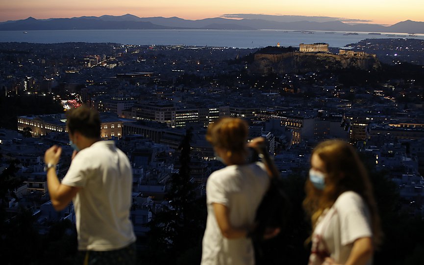 A family of tourists wearing face masks against the spread of the coronavirus, walk on Lycabettus hill as the Parthenon temple is illuminated atop of the Acropolis hill, background right during the sunset in Athens, Monday, Oct. 26, 2020. (AP Photo/Thanassis Stavrakis)
