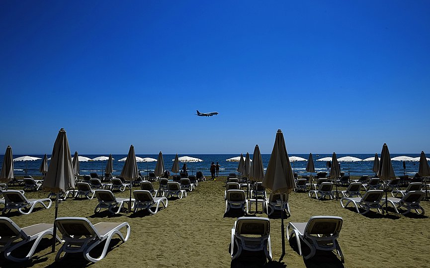 People stand by the sun-beds and umbrellas as an aircraft prepares for landing at Larnaca international airport, at Makenzi beach in southern city of Larnaca, Cyprus, Tuesday, May 18, 2021. (AP Photo/Petros Karadjias)